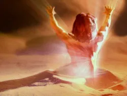 Person kneeling in sand with raised arms, glowing light shining around them.