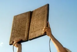 Two hands holding large stone tablets with inscriptions against a blue sky.